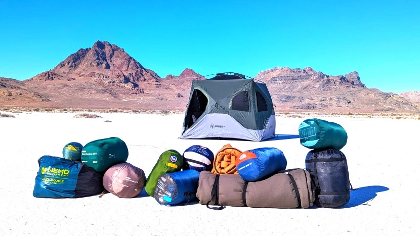 Various sleeping bags displayed in outdoor mountain camping setting showing different temperature ratings and styles